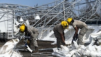 Troops in hard hats clean up debris on the ground near mangled metal structures.