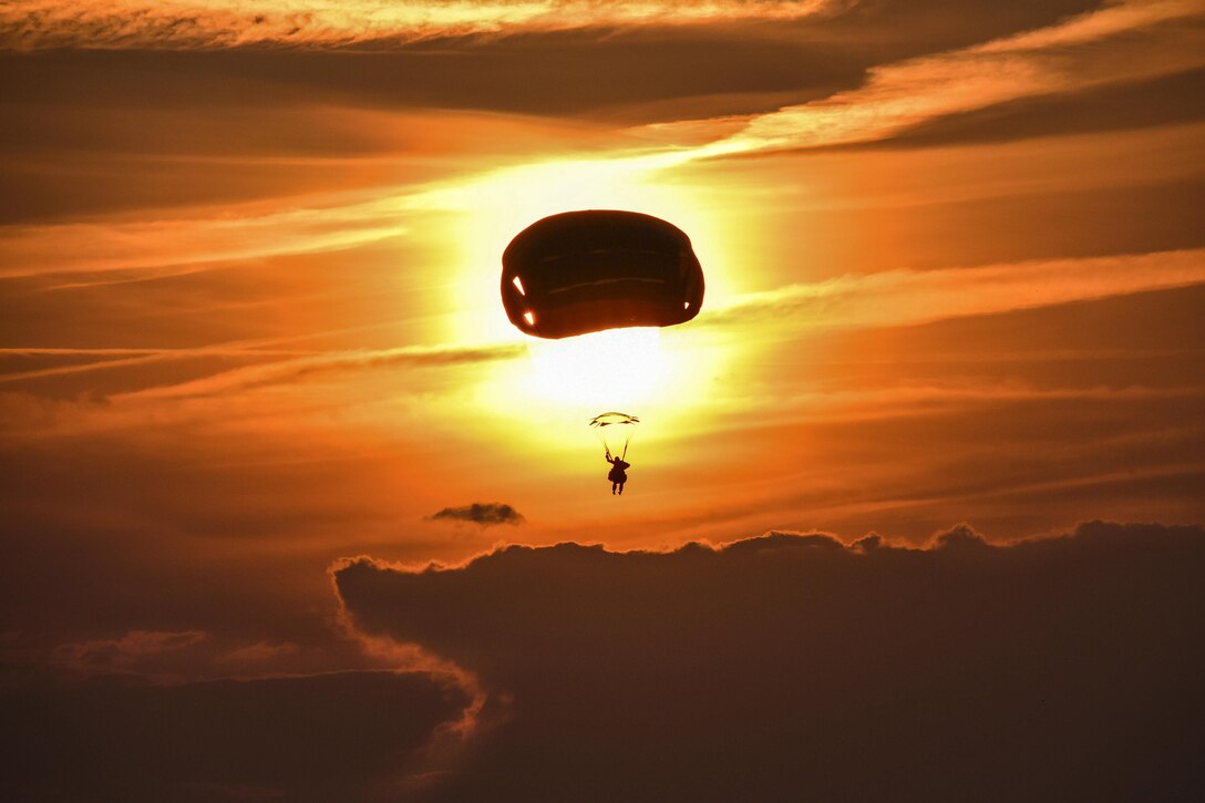 A soldier, shown in silhouette, parachutes toward the ground against an orange sky.