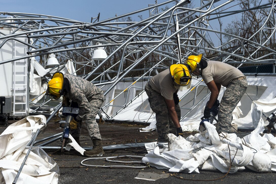 Troops in hard hats clean up debris on the ground near mangled metal structures.