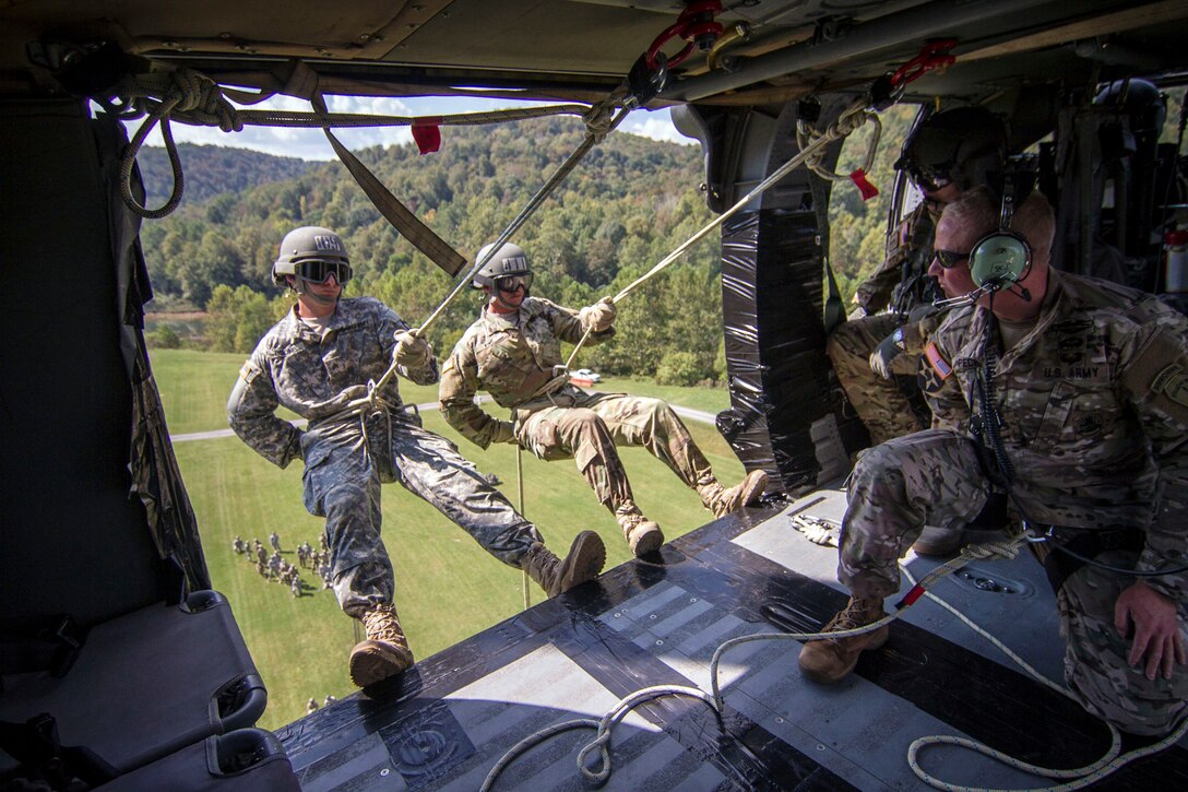 Two soldiers suspended by ropes lean out the open door of an in-flight helicopter.