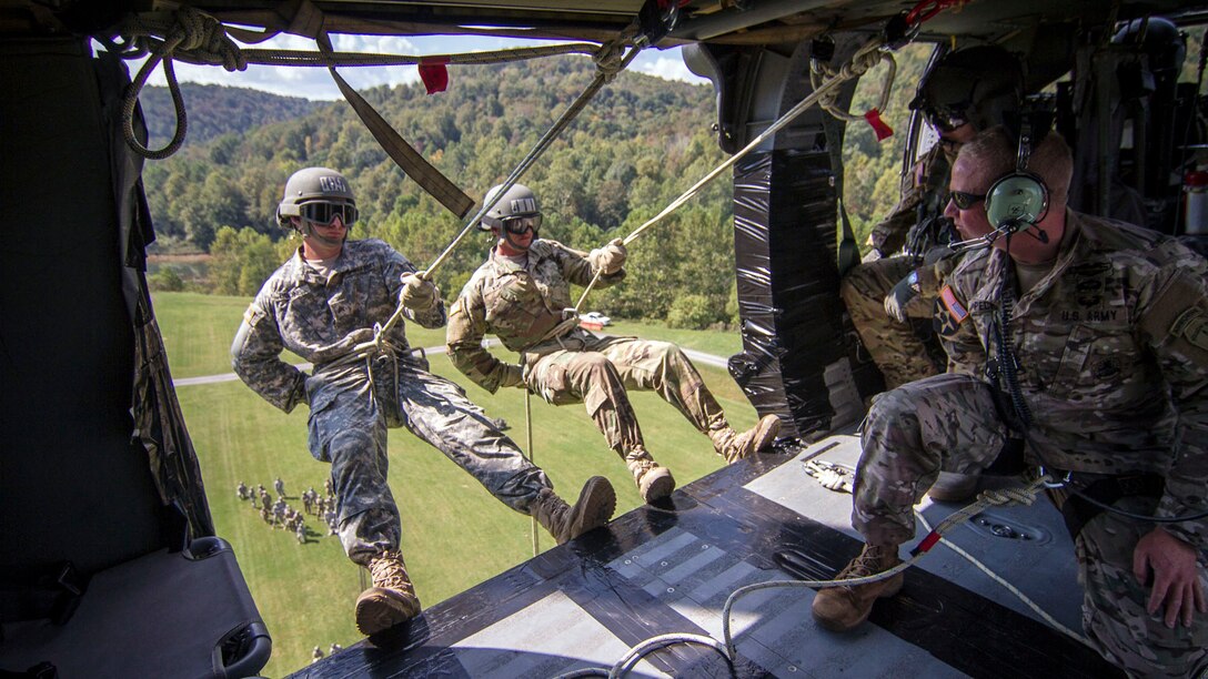Two soldiers suspended by ropes lean out the open door of an in-flight helicopter.