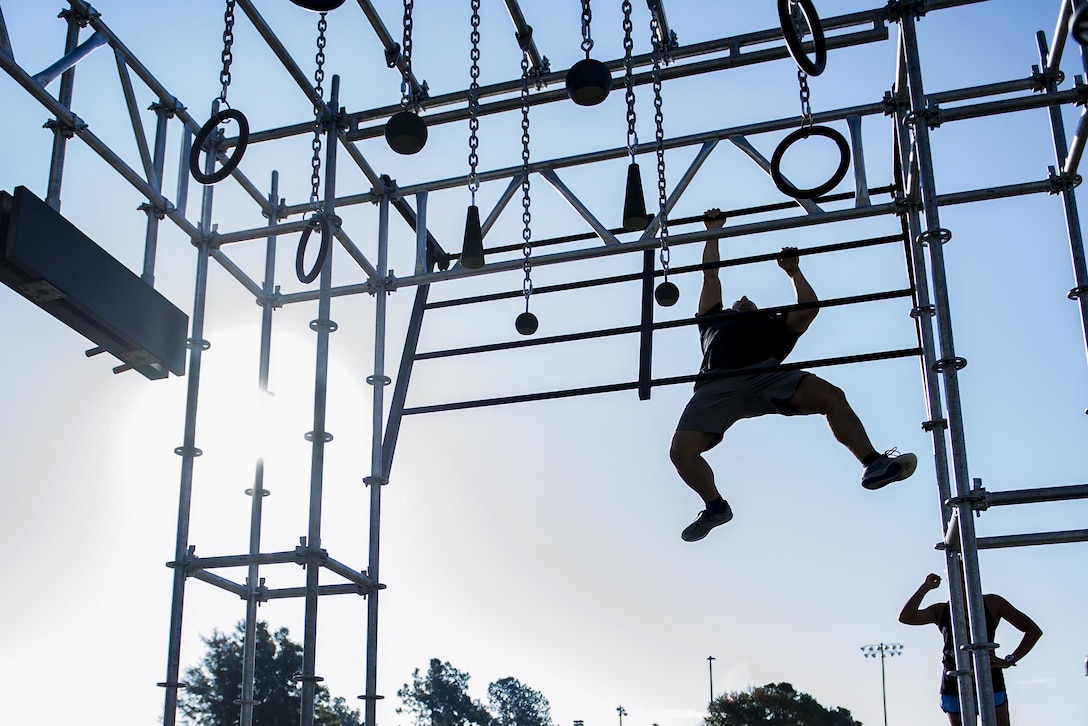 An airman, shown in silhouette, uses his arms to move across metal bars on an obstacle.