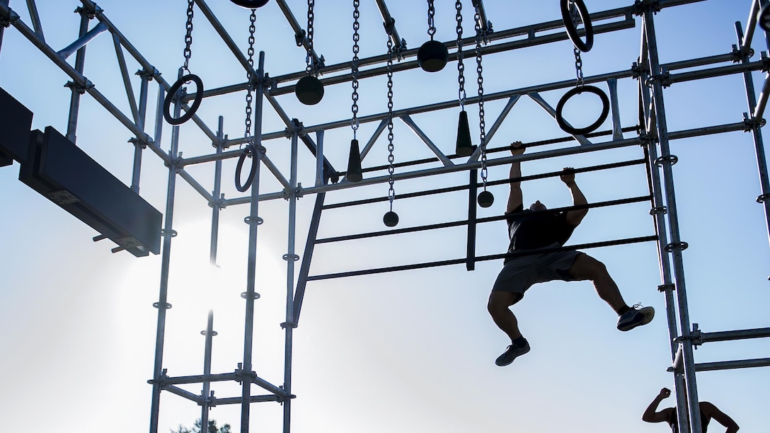 An airman, shown in silhouette, uses his arms to move across metal bars on an obstacle.