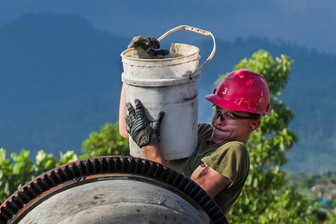 A Marine standing at the mouth of a cement mixer holds up a large bucket.