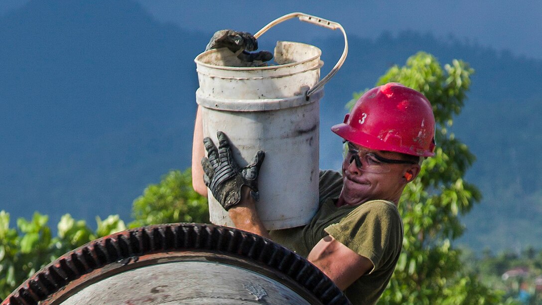 A Marine standing at the mouth of a cement mixer holds up a large bucket.