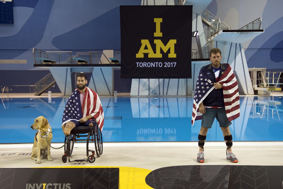 Two wounded warriors draped in the american flag take the stage, one in a wheelchair while holding the leash to a dog,