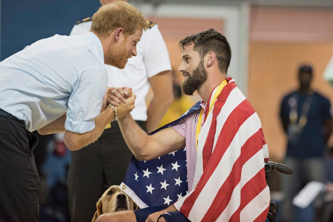 Prince harry and a wounded warrior in a wheelchair shake hands.