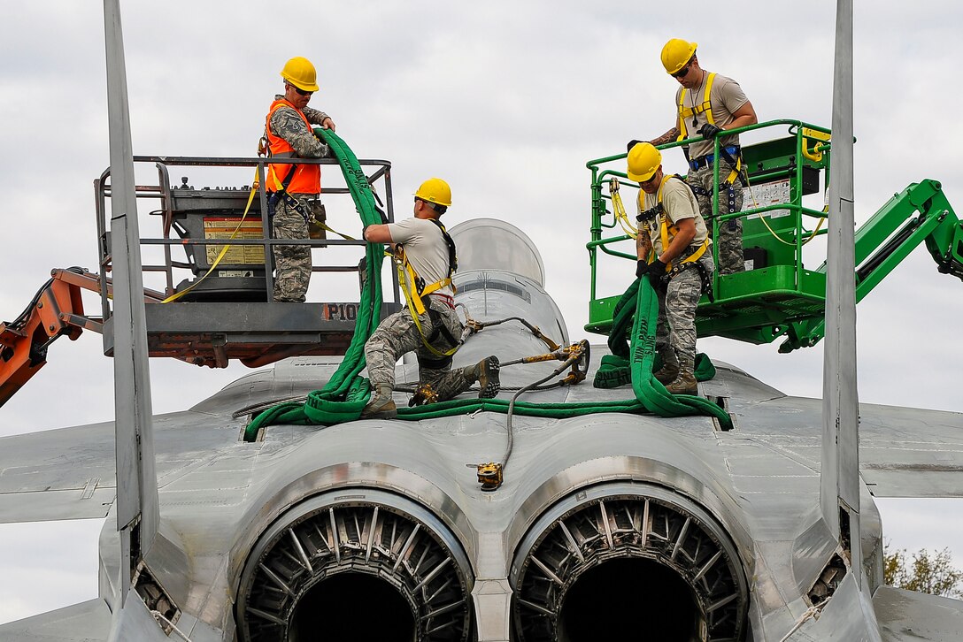 Four airmen in yellow hardhats work atop a stationary fighter jet.