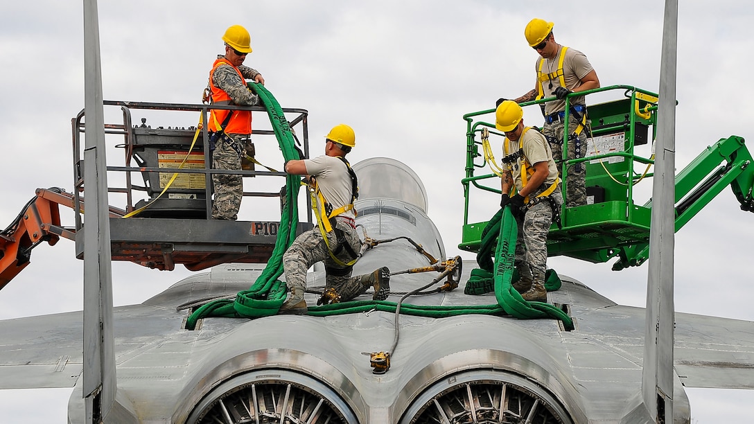 Four airmen in yellow hardhats work atop a stationary fighter jet.