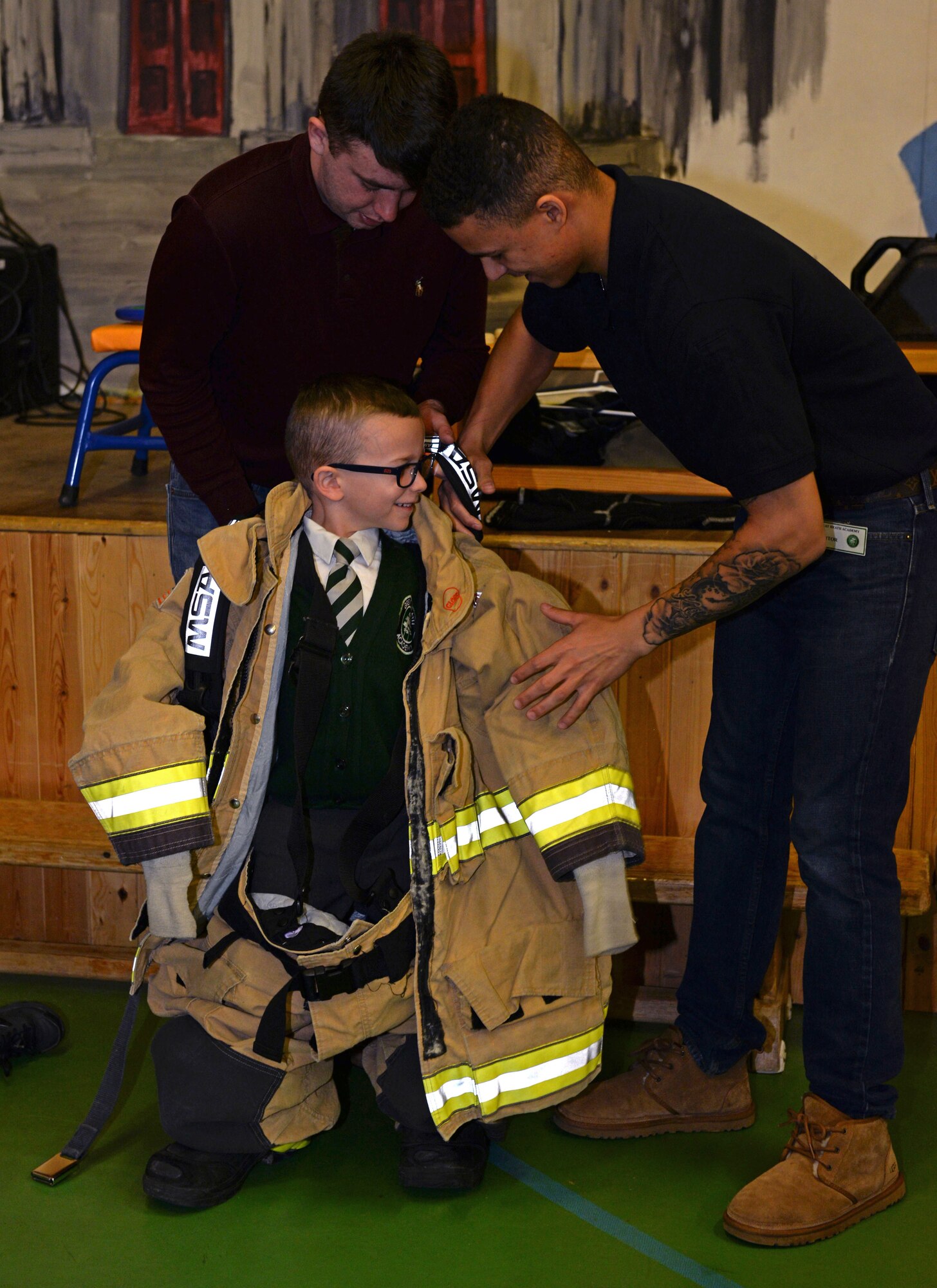U. S. Air Force Airmen 1st Class Levi Brakenwagon and Isaiah Gwaltney, both 100th Civil Engineer Squadron firefighters, help a primary school student as he tries on a set of bunkers during a school visit at Great Heath Academy in Mildenhall, England, March 27, 2018. The firefighters participate in several volunteer events throughout the year at schools in the local community, focusing on educating children on fire safety. (U.S. Air Force photo by Airman 1st Class Alexandria Lee)