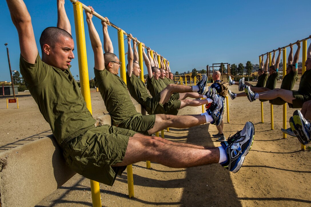 Marine recruits hang from a yellow bar and lift their legs parallel to the ground.