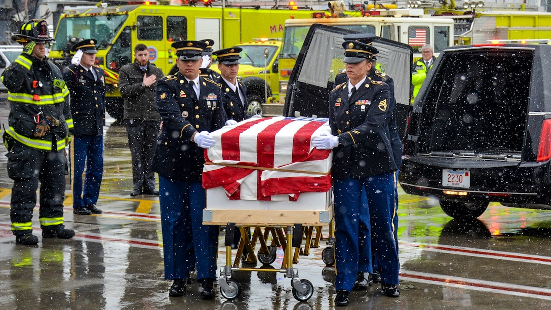 Soldiers carry a flag draped casket on a snowy flightline as personnel nearby salute.