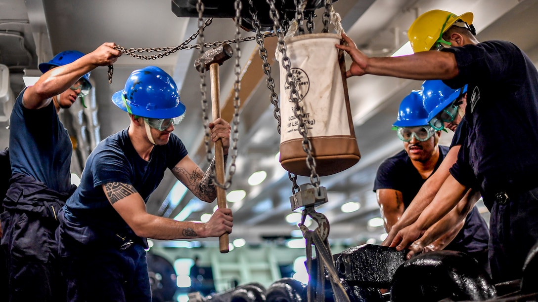 A sailor wields a mallet as others manipulate an anchor chain.