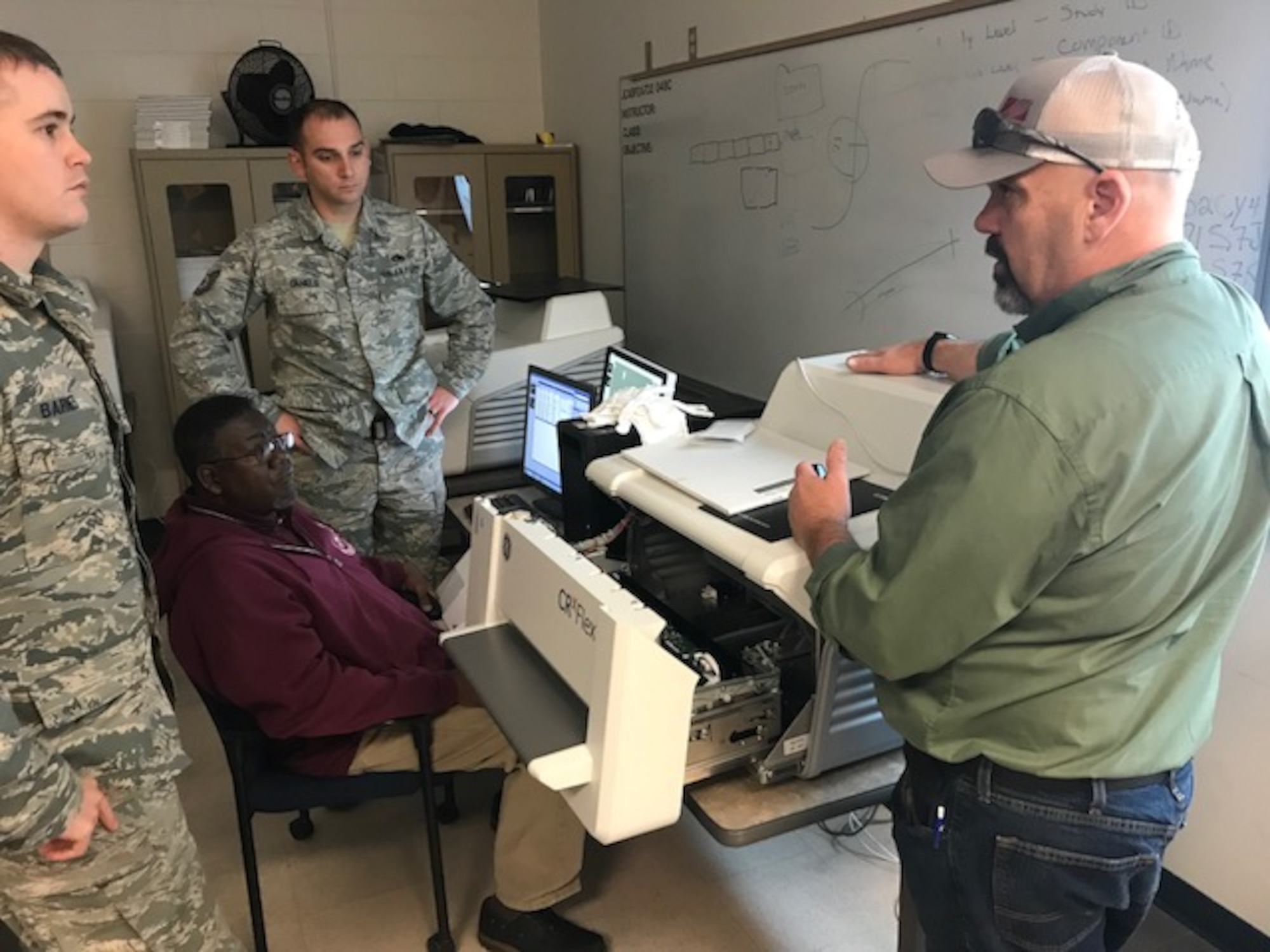 Kevin McClain, contract nondestructive testing instructor, explains how to clear a jammed image scanner from a computed radiography system to visiting Nondestructive Inspection Instructor, Tech. Sgt. Matthew Barnes (left); Tech. Sgt. Clifford Daniels, Military NDI Instructor supervisor (rear); and Bobby Britton, Civilian NDI Instructor supervisor (seated) during instructor qualification training held at the 359th Training Squadron Detachment One at Naval Air Station Pensacola, Florida Jan. 24, 2018. New digital technology will replace the X-ray film used by NDI technicians over the next several years and impact every airframe the Air Force flies, saving money, reducing hazardous materials and improving operational capabilities. (Courtesy photo)