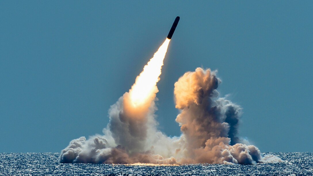 A missile fires out of the ocean at a diagonal trajectory, causing smoke to waft up from the blue water.