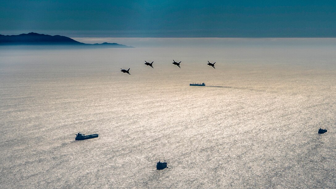 Four aircraft fly over ships in still waters.