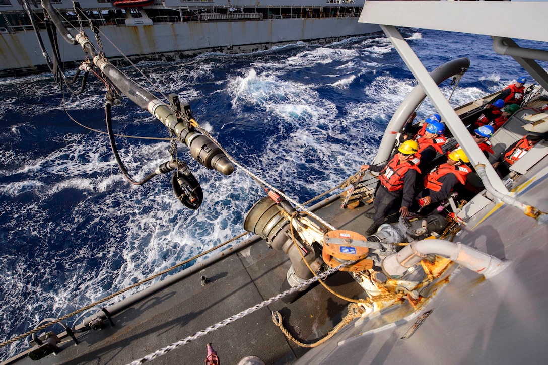 Sailors on a ship observe as a fuel line from a nearby ship extends to connect with the intake on theirs.