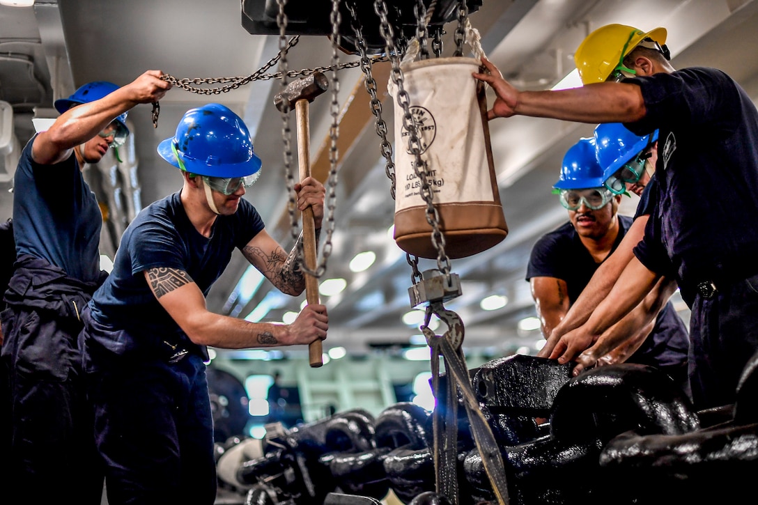 A sailor wields a mallet as others manipulate an anchor chain.