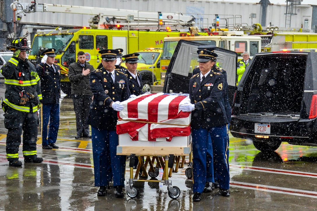 Soldiers carry a flag draped casket on a snowy flightline as personnel nearby salute.