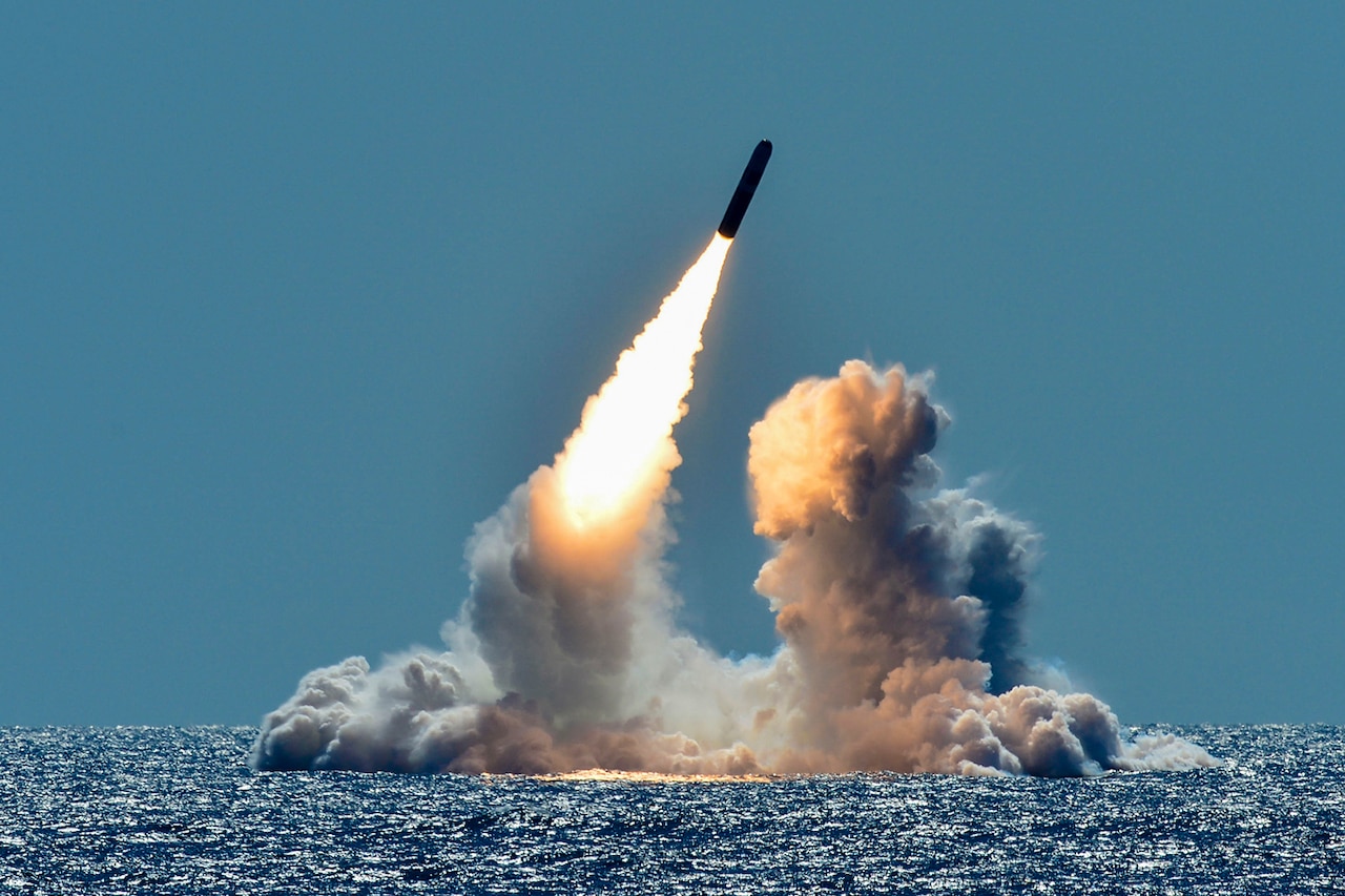 A missile fires out of the ocean at a diagonal trajectory, causing smoke to waft up from the blue water.