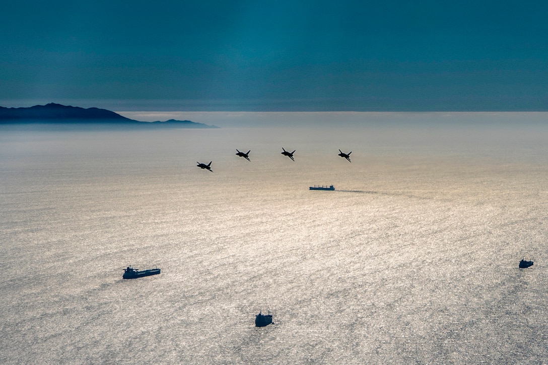 Four aircraft fly over ships in still waters.