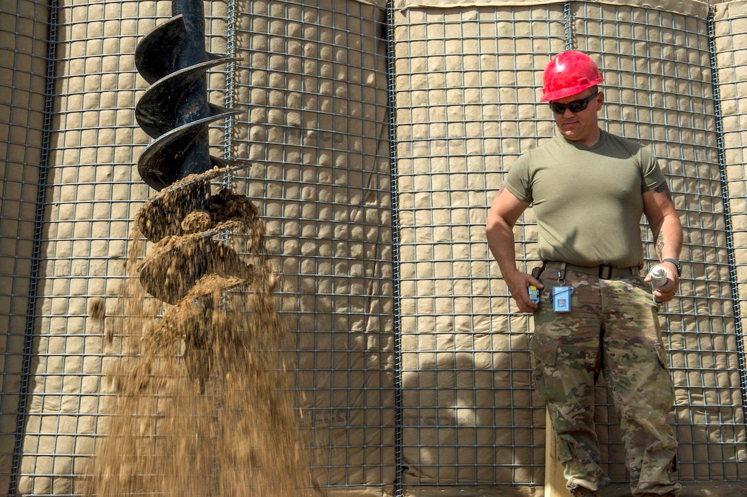 A soldier watches as another soldier operates an auger dirt driller.