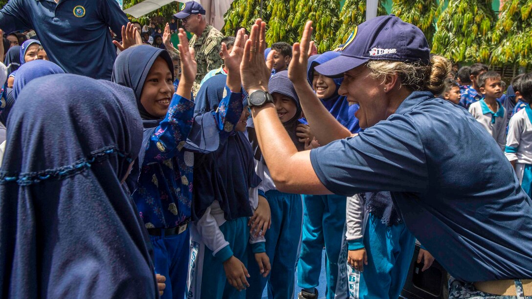 A sailor holds both hands up and smiles as a schoolgirl does the same.