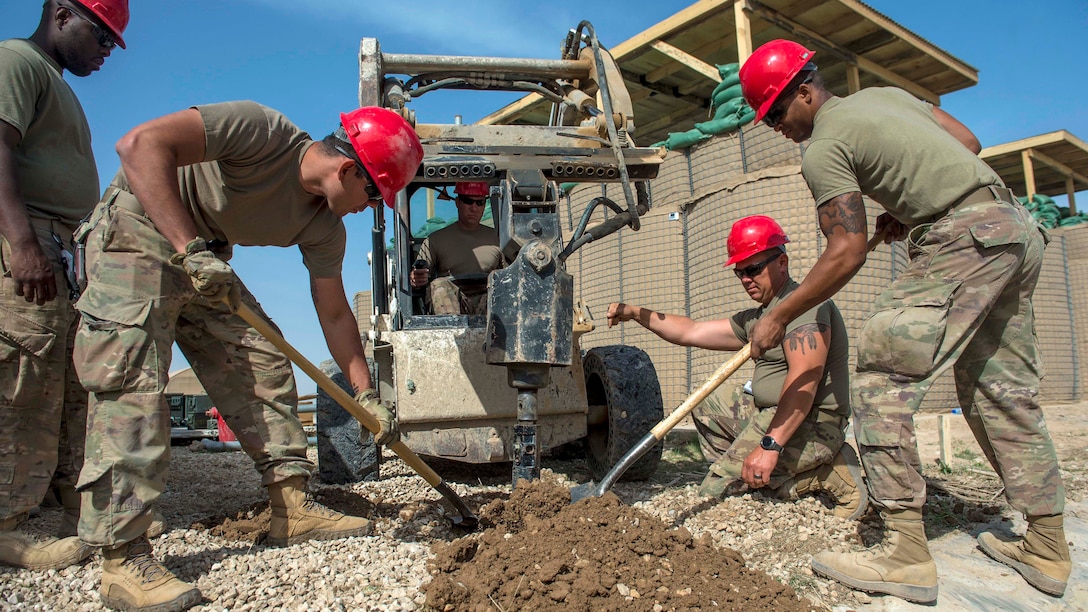 Soldiers in red hard hats dig in a patch of dirt.