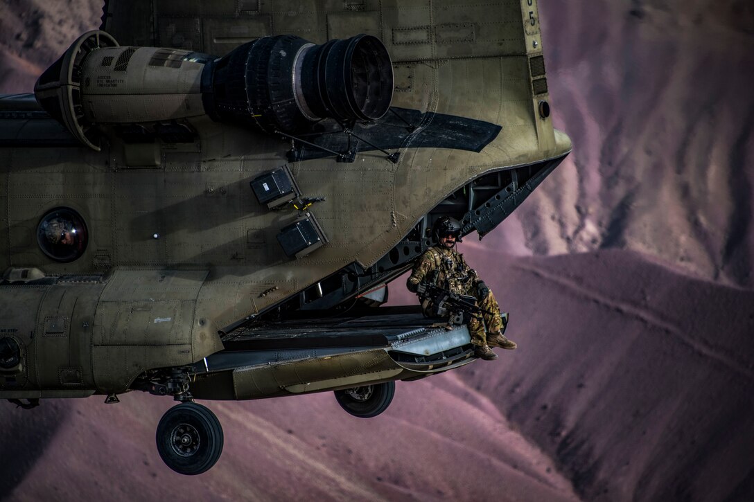 A soldier sits in the back of an open helicopter flying over dramatic purplish-brown hills.