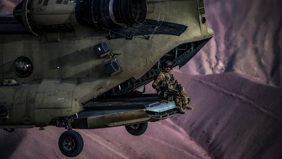 A soldier sits in the back of an open helicopter flying over dramatic purplish-brown hills.