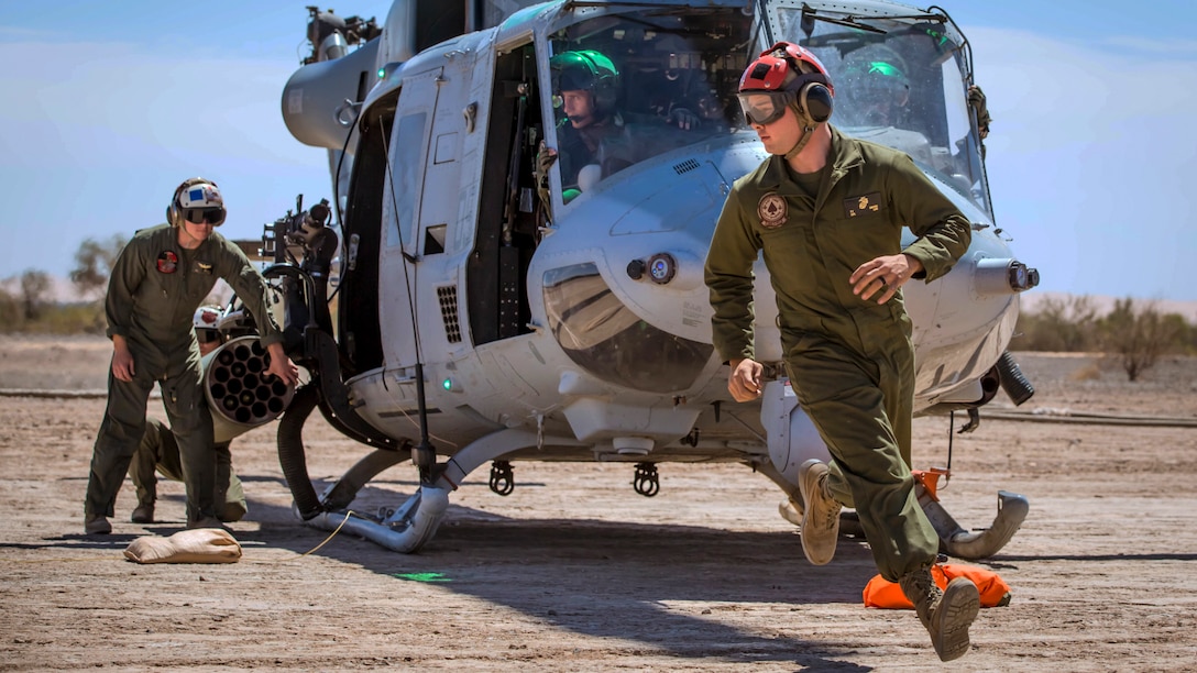 A Marine rushes in front of a helicopter in a dirt field as others work in and around the aircraft.