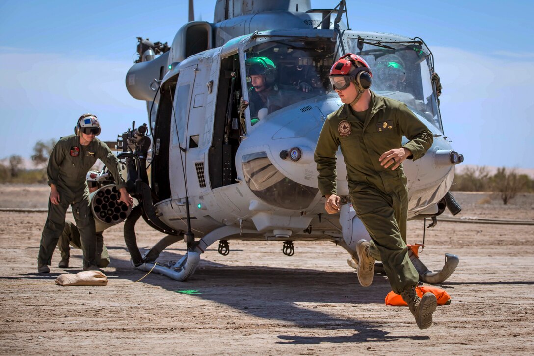 A Marine rushes in front of a helicopter in a dirt field as others work in and around the aircraft.