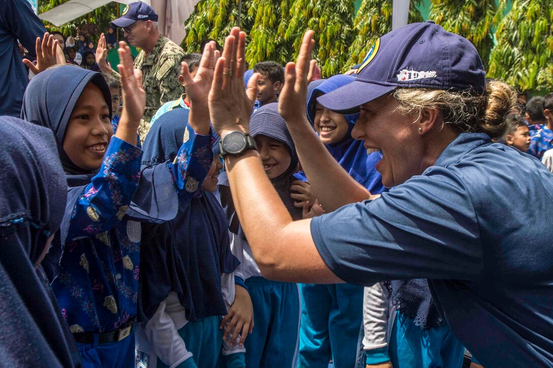 A sailor holds both hands up and smiles as a schoolgirl does the same.