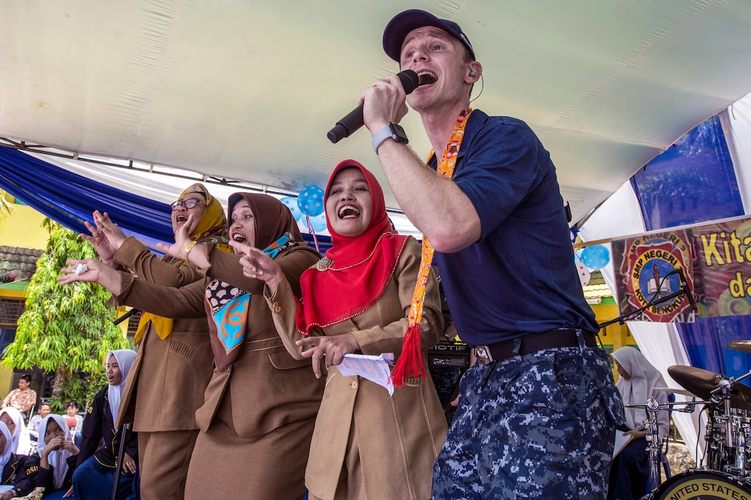 A sailor sings into a microphone as three women dance and sing alone beside him.