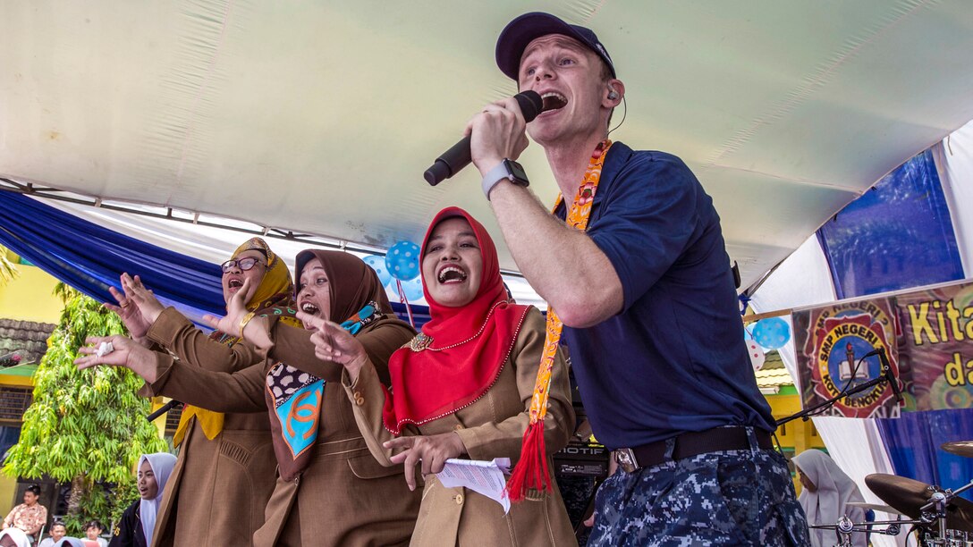A sailor sings into a microphone as three women dance and sing alone beside him.