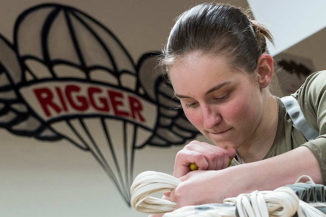 A soldier manipulates rope in a room with a wall mural of a winged parachute that says "RIGGER" in red letters.