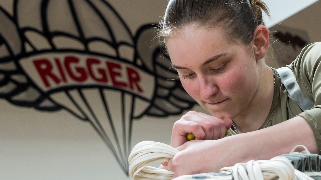 A soldier manipulates rope in a room with a wall mural of a winged parachute that says "RIGGER" in red letters.