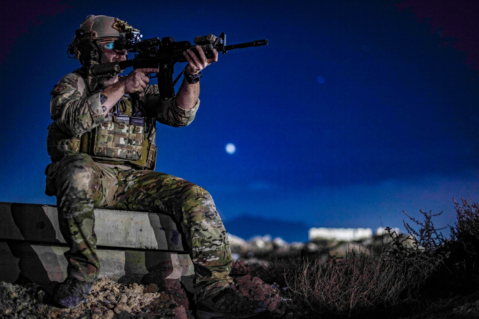 An airman sits on a ledge and aims a rifle against a deep blue, moonlit sky.