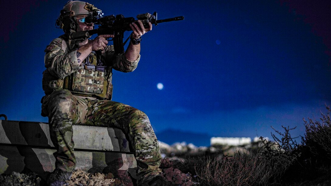 An airman sits on a ledge and aims a rifle against a deep blue, moonlit sky.