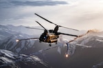 A soldier sits in the back of an open helicopter flying over dramatic purplish-brown hills.