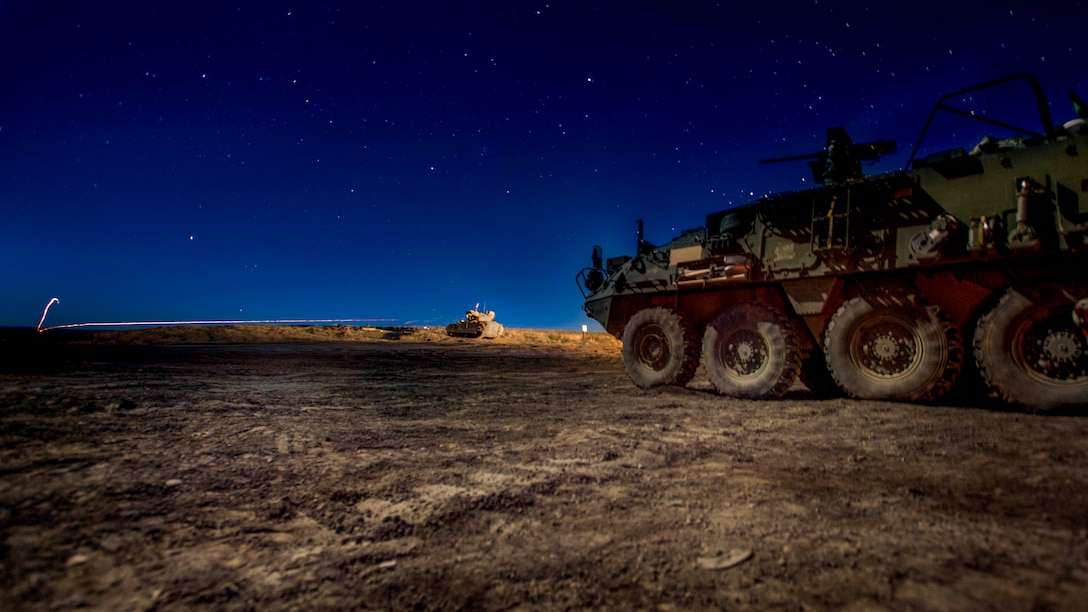 A fighting vehicle fires a lighted projectile in the distance as another sits in the foreground in a field against a deep blue sky.