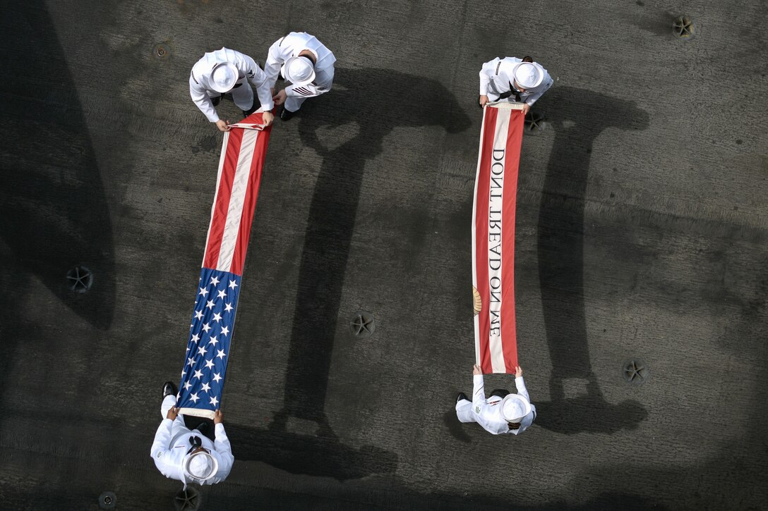 Five sailors, seen from overhead, fold two red and white flags on a dark grey flight deck.
