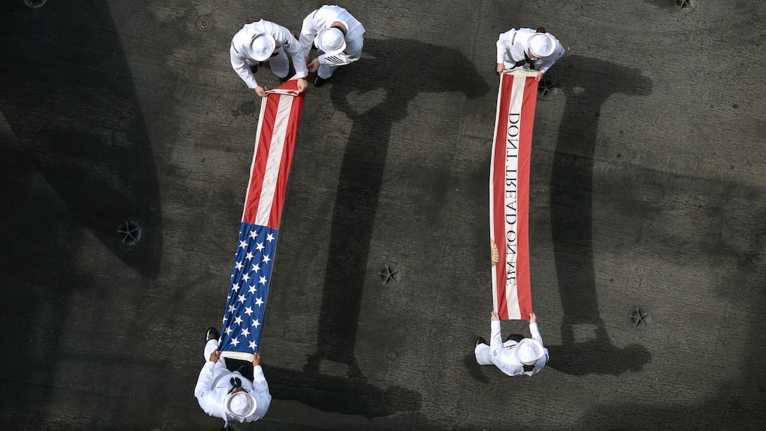 Five sailors, seen from overhead, fold two red and white flags on a dark grey flight deck.