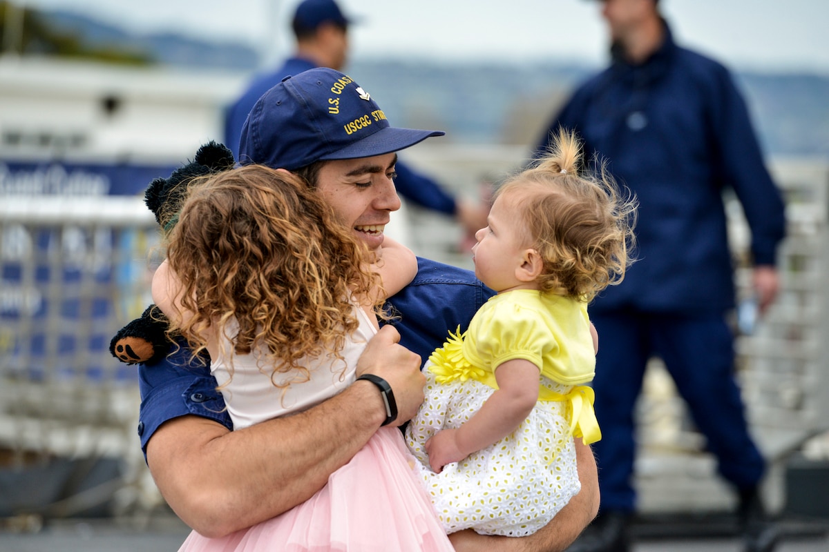 A Coast Guardsman embraces two little girls, one of whom holds a teddy bear, on a pier.