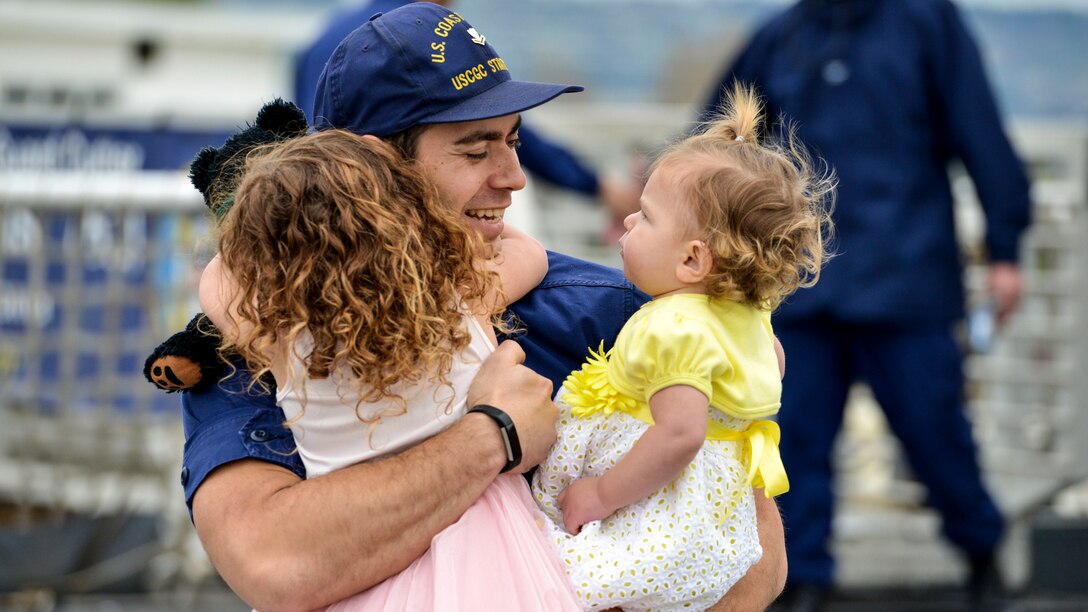 A Coast Guardsman embraces two little girls, one of whom holds a teddy bear, on a pier.