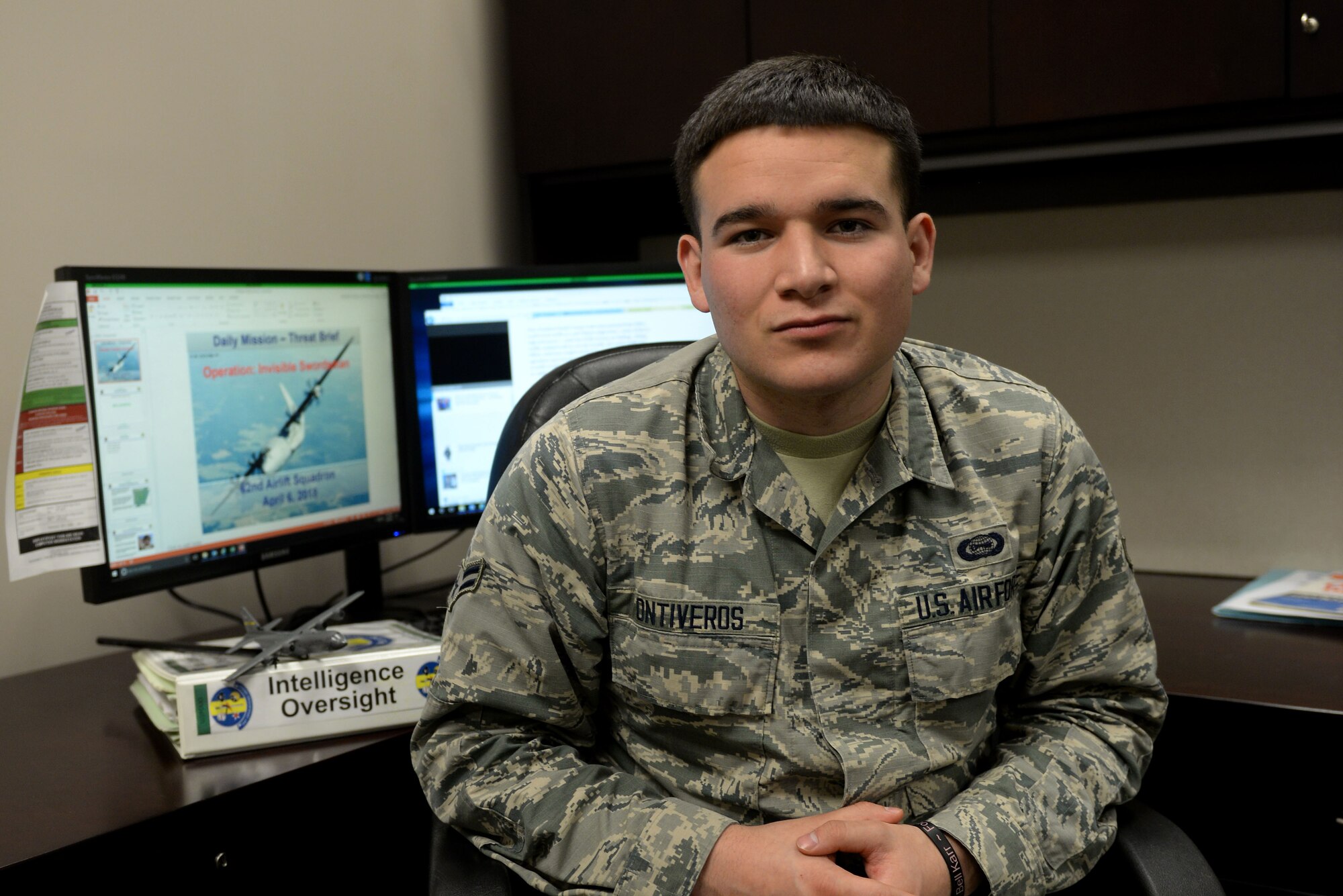 A young man sits in a chair while wearing the Airman Battle Uniform with two computer monitors behind him.
