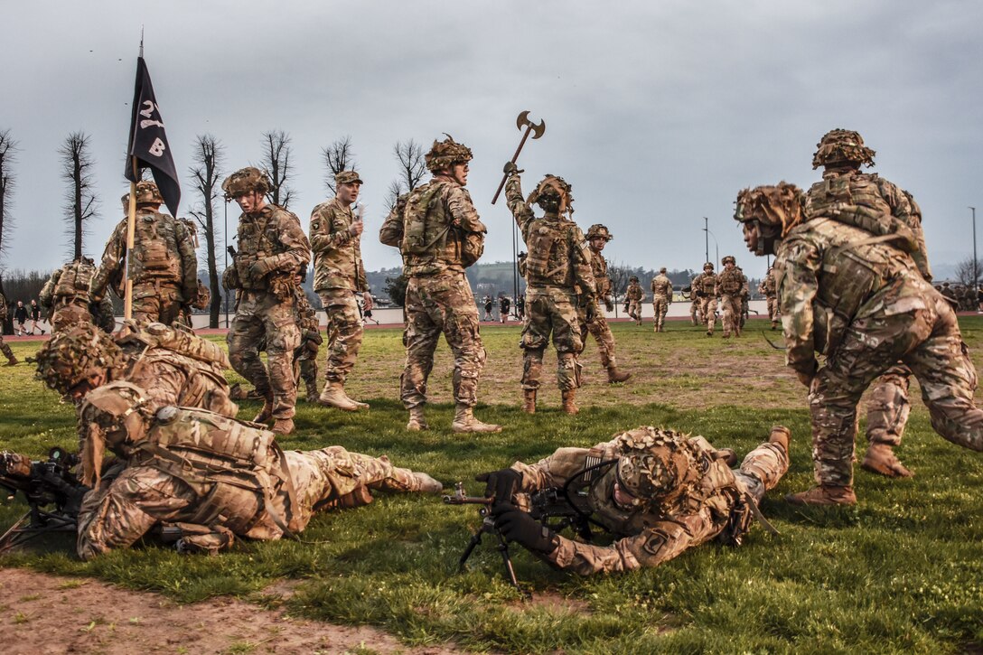 A soldier holds up a medieval-style battle ax to mark the finish line for marchers in a field as others gather around him.