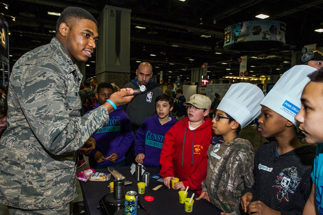 An airman standing at a table holds up a piece of a metal while talking to students gathered around.