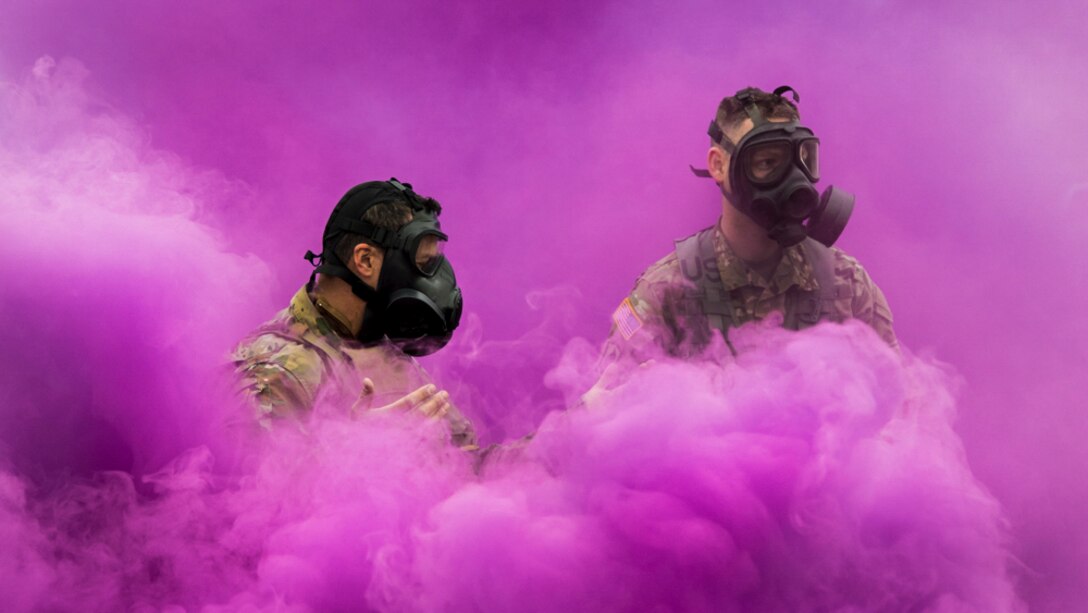 Two soldiers wearing gas masks stand in a thick cloud of mauve-colored smoke.