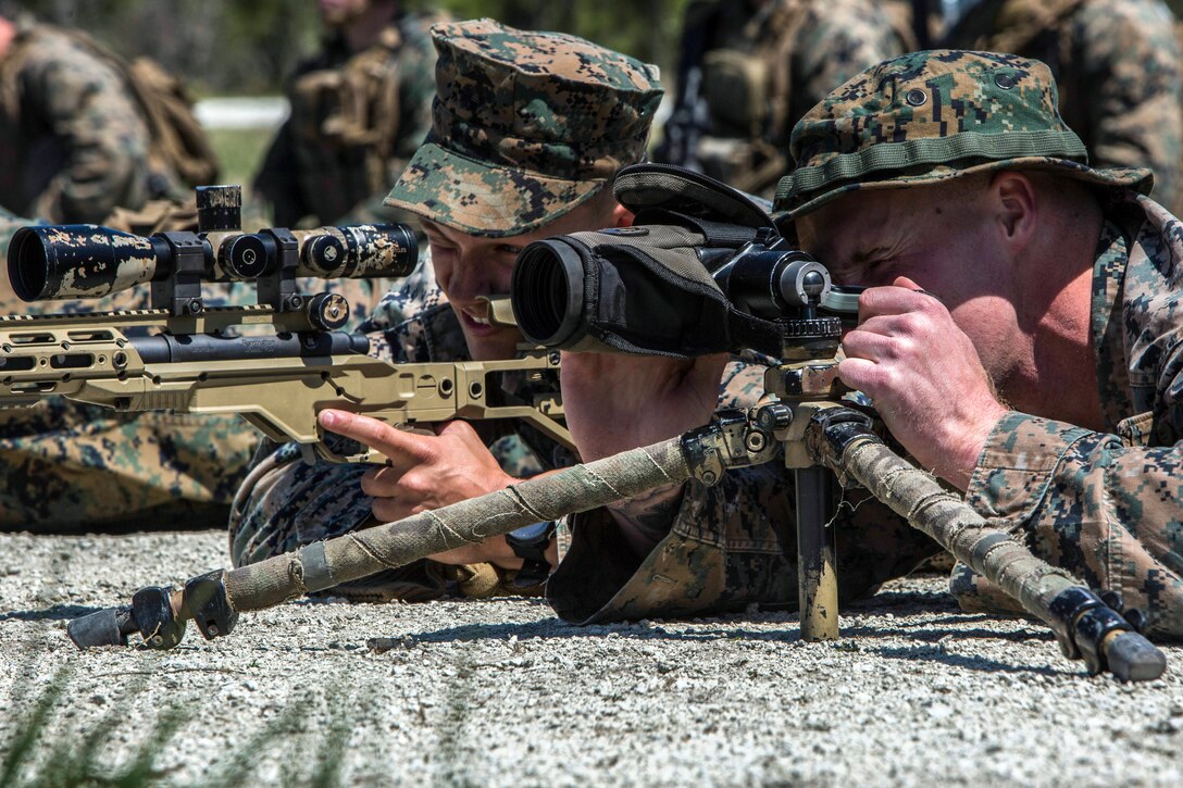 Two Marines lying behind weapons squint while looking through weapon scopes.